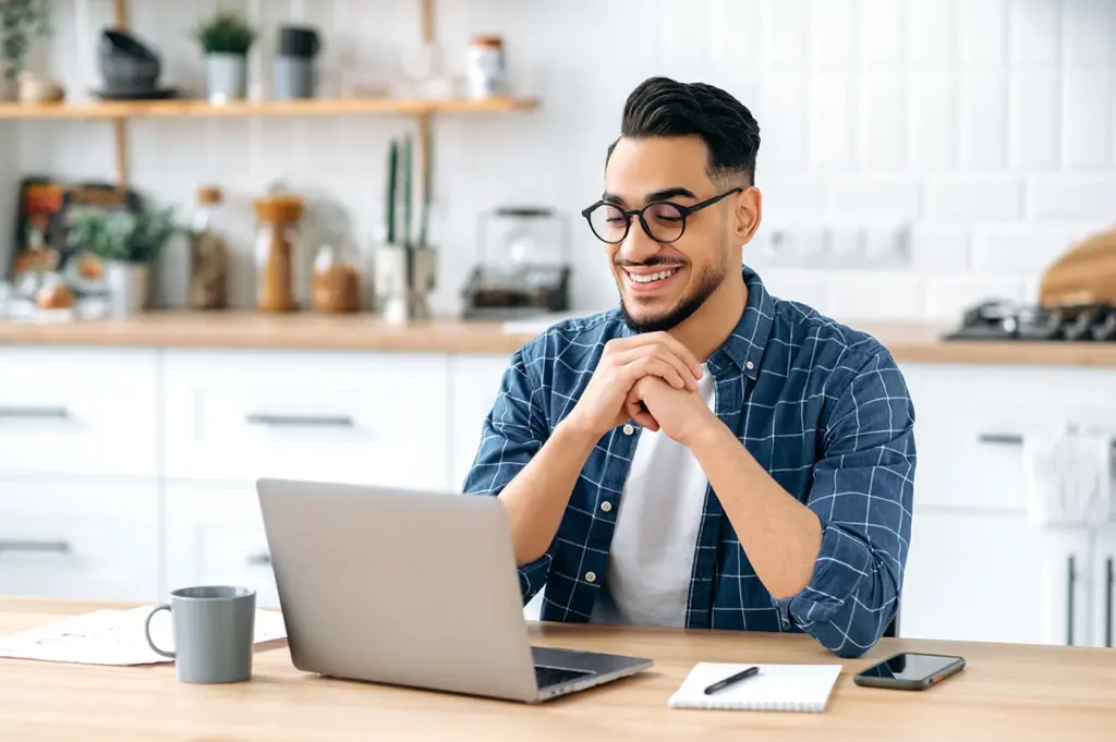 Smiling man wearing glasses and a checkered shirt sits at a kitchen table using a laptop, with a notebook, pen, coffee mug, and smartphone beside him.