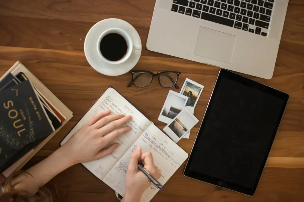 A top-down view of a wooden desk with an open notebook, a person writing with a pen, a cup of black coffee, a laptop, a tablet, eyeglasses, several instant photos, and a stack of books.