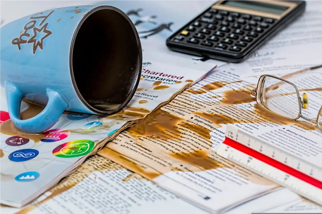 A spilled blue coffee mug lies on its side, soaking printed documents, a pair of eyeglasses, a calculator, and a ruler with coffee, creating a messy and chaotic workspace scene.