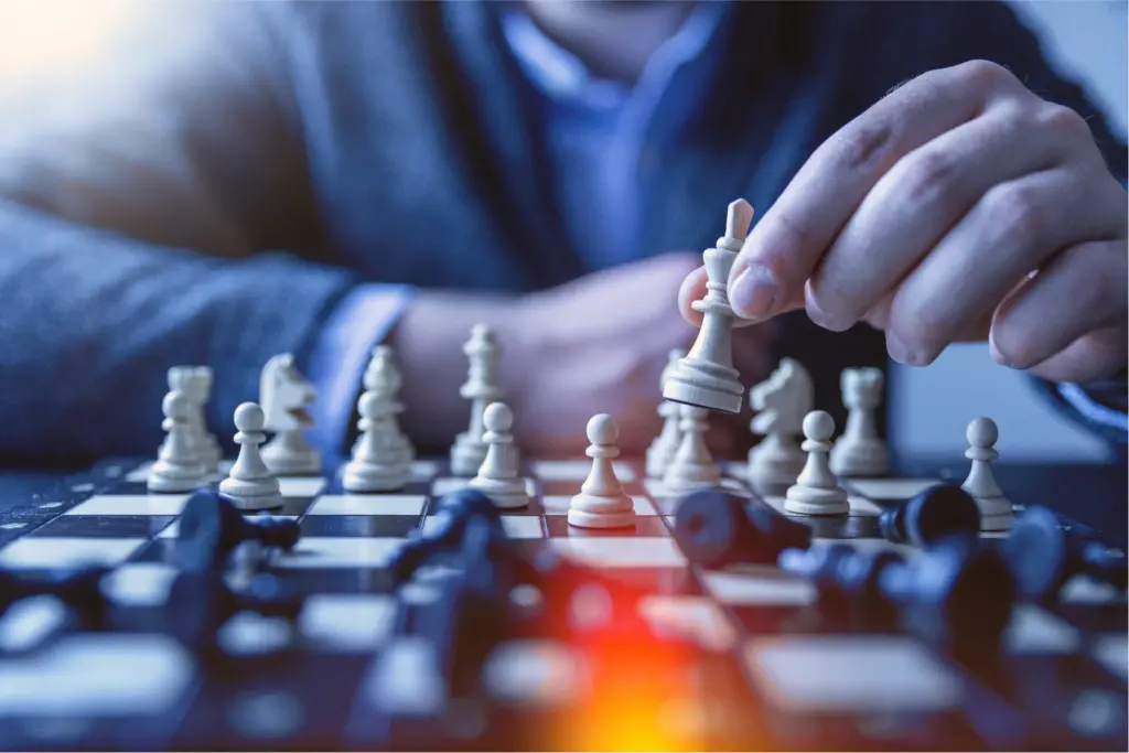 Close-up of a person’s hand moving a white king piece during a chess game, with scattered black pieces and a blurred background suggesting strategic thinking.