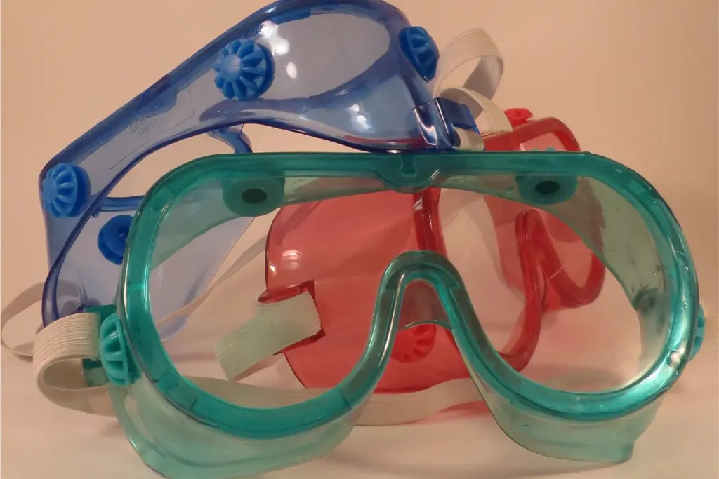 Three pairs of translucent safety goggles in teal, red, and blue, stacked together with white elastic straps against a neutral background.