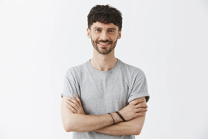 A smiling man with short dark hair and a beard stands with his arms crossed, wearing a light gray t-shirt against a plain white background.