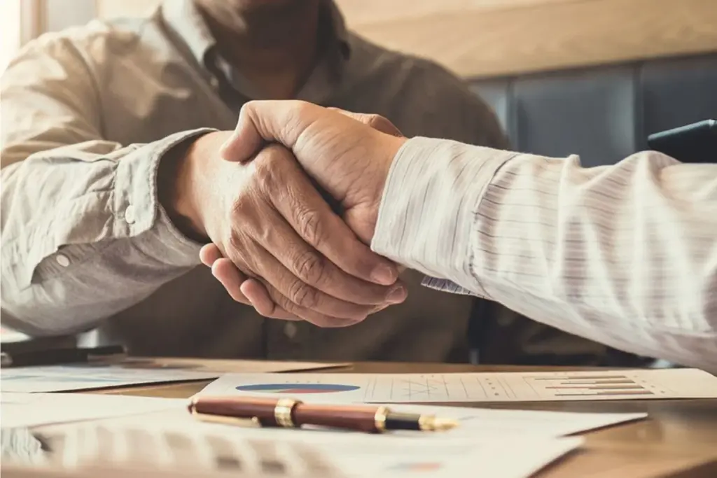 Two people shaking hands across a table with business documents and a pen, symbolizing agreement or a successful negotiation.