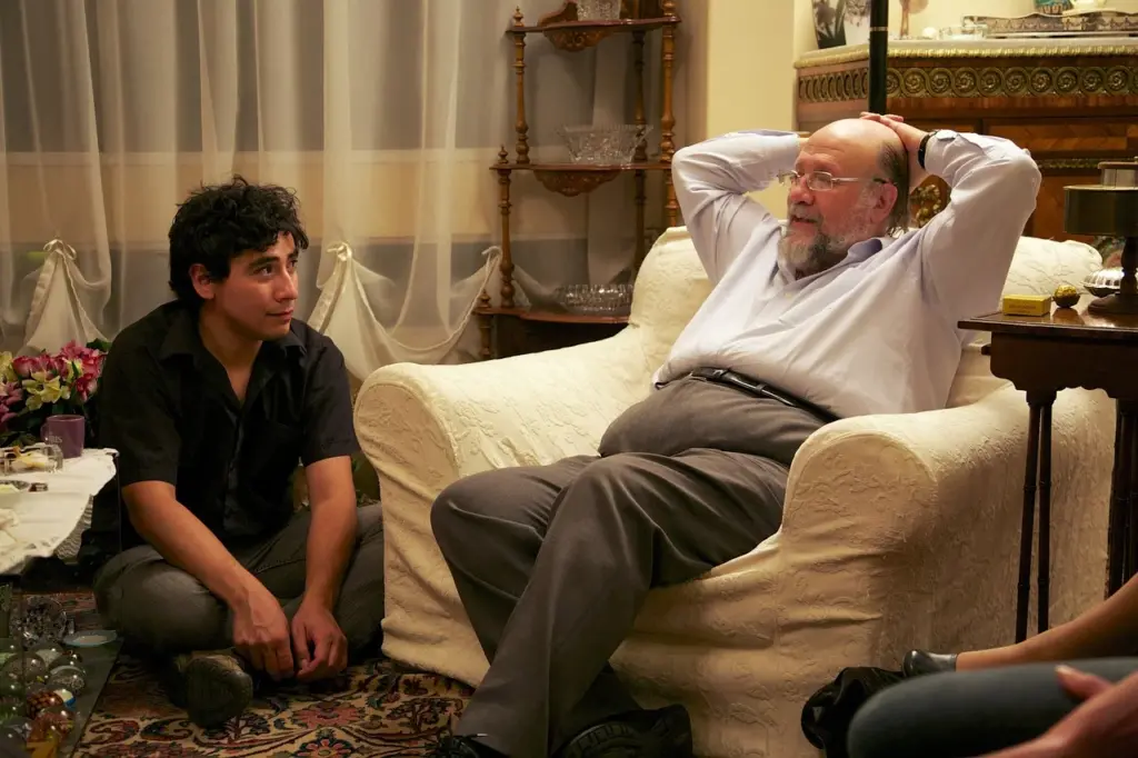 Older man sitting relaxed in an armchair with hands behind his head, speaking to a younger man seated on the floor listening attentively, suggesting a mentor-mentee conversation in a cozy, well-decorated room.