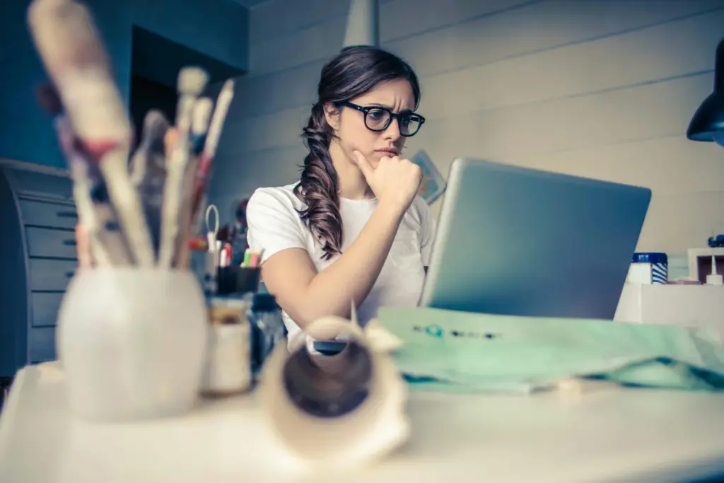 A young woman wearing glasses and a white shirt sits at a desk, looking thoughtfully at a laptop screen with her hand on her chin. The foreground shows a blurred collection of paintbrushes and art supplies, suggesting a creative workspace.