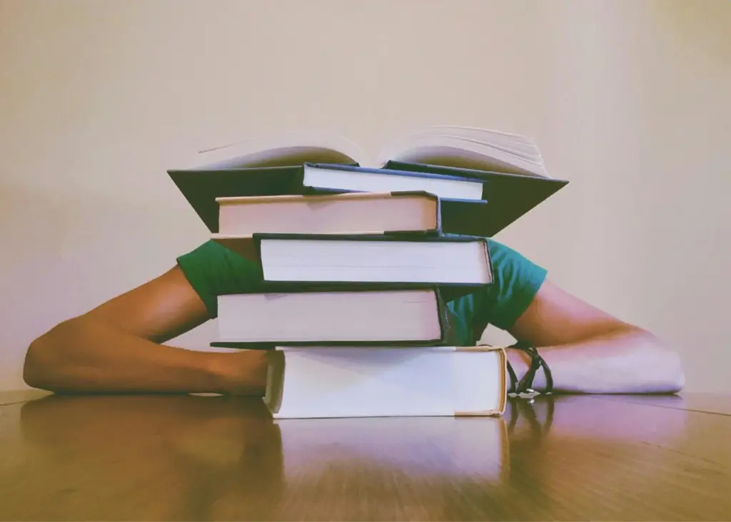 A person in a green shirt sits at a table with their head resting behind a tall stack of books, one of which is open on top. Only their arms are visible, suggesting exhaustion or overwhelm from studying.