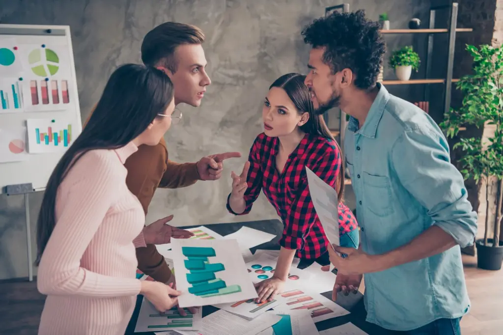 Four young adults gather around a table covered in papers and charts, engaged in an intense discussion or debate, with some gesturing and pointing. A whiteboard with graphs is in the background.