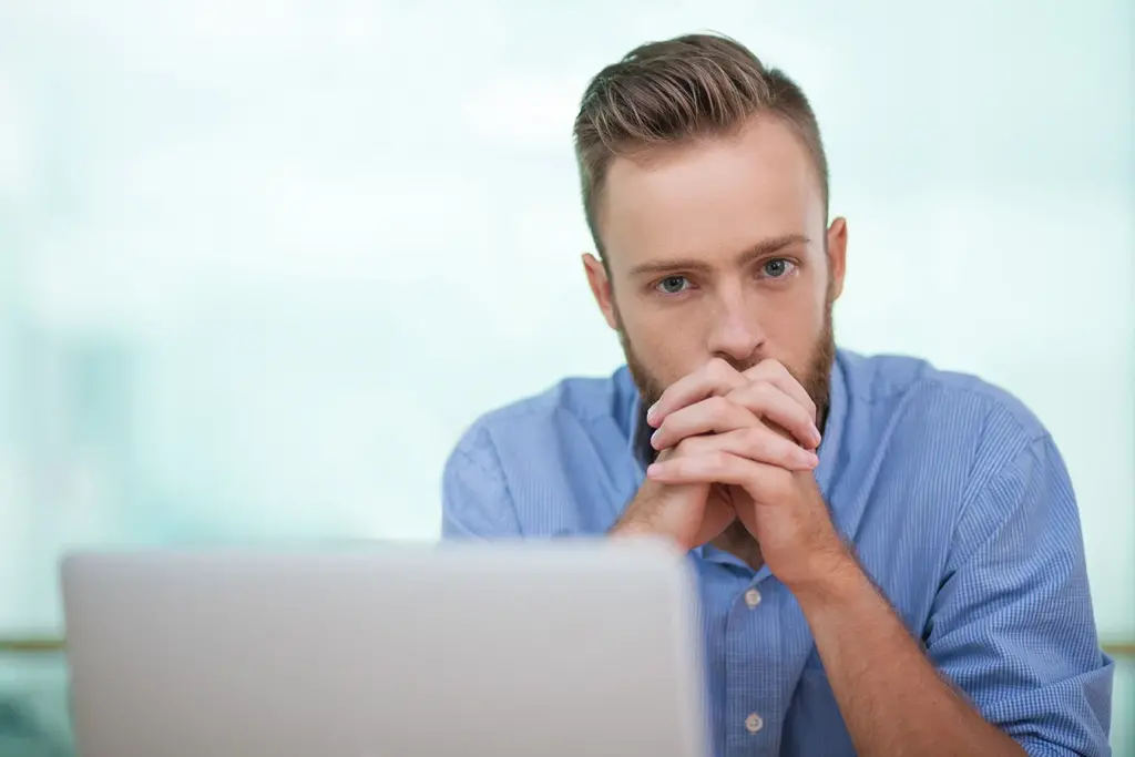 A man in a blue shirt sits behind a laptop, resting his chin on his clasped hands, appearing deep in thought or contemplation.