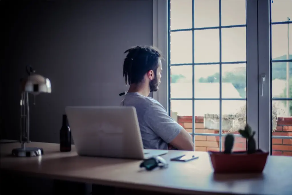 A man with dreadlocks sits at a desk with a laptop, looking thoughtfully out a window with grid panes; a dimly lit room contains a lamp, a beer bottle, and a small potted plant.