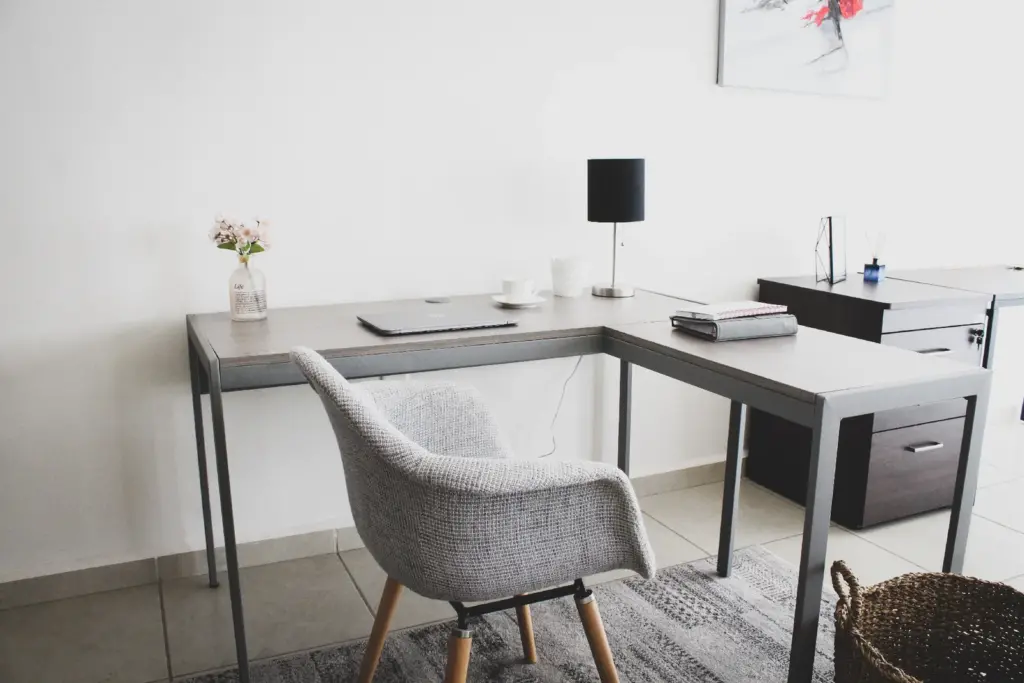 Modern minimalist home office with a gray L-shaped desk, upholstered chair, laptop, lamp, notebook, and decorative items like a small vase with flowers and a woven basket on the floor.