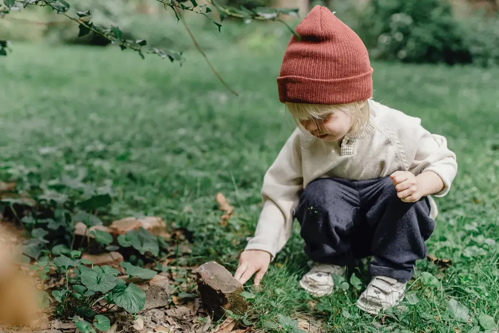 A young child in a red knit hat and cream sweater crouches on grass, reaching down to touch a brick among plants and leaves, appearing curious and engaged in outdoor exploration.