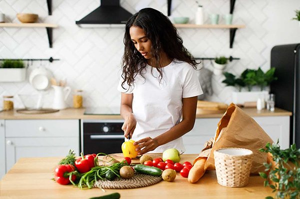 A woman in a white shirt stands in a modern kitchen, slicing a yellow bell pepper on a wooden counter surrounded by fresh vegetables and a paper grocery bag.